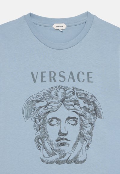 Ανοιχτό μπλε μπλούζα με το λογότυπο Versace πάνω από μια λεπτομερή μαύρη γραμμική σχεδίαση του κεφαλιού της Μέδουσας μπροστά.