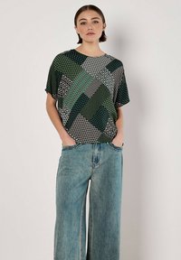 Kurzärmliges Oberteil mit grünen, schwarzen und weißen geometrischen Mustern, kombiniert mit weit geschnittenen, ausgebleichten blauen Jeans. Glattes Material und entspannte Passform.