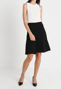 Top blanco sin mangas con un patrón texturizado y una falda negra de corte A. Combinado con tacones negros de punta. Tejido suave y ajustado.