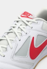 Balti sportiniai bateliai, pagaminti iš tinklelio ir odos, su raudonu Nike logotipu, pilkais akcentais ir plokščiomis baltomis raištelėmis.