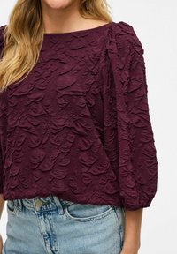 Žena nese texturovanou, tmavě fialovou blůzu s nabíranými rukávy v kombinaci s světle modrými džínami.