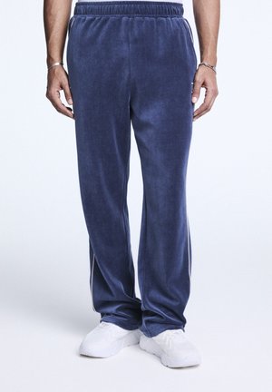 Homme portant un pantalon en velours bleu foncé ample et des baskets blanches, debout, les mains le long du corps, sur un fond uni.