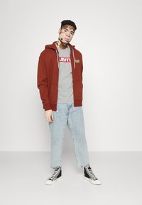 Sudadera con capucha y cremallera en color naranja quemado, con puños de canalé, combinada con una camiseta gris de Levi's, jeans cortos de mezclilla azul claro y zapatillas altas negras.