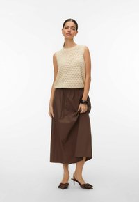 Femme debout portant un haut beige en tricot sans manches, une jupe midi marron, des talons marron à pois, un bracelet noir et une bague à la main.