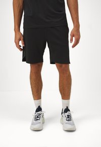 Pantalones cortos deportivos negros con una textura suave, diseño hasta la rodilla y acentos de panel lateral en contraste. Combinados con zapatillas deportivas blancas y calcetines.