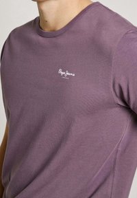 Lilla bomuld T-shirt med rund hals, korte ærmer og hvidt logo på venstre brystområde. Blød tekstur med afslappet pasform.