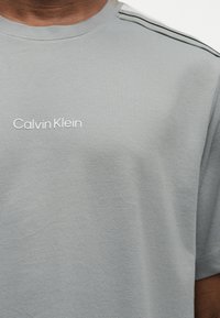 Ανοιχτό γκρι μπλουζάκι με το λογότυπο Calvin Klein στο στήθος, φορεμένο από άτομο, με μαύρες διπλές ρίγες στον ώμο.