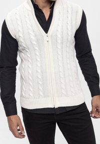 Kabelgebreid, crème mouwloos vest met een ritssluiting aan de voorkant; heeft een gestructureerd patroon en een geribde zoom; gedragen over een zwart shirt met lange mouwen.