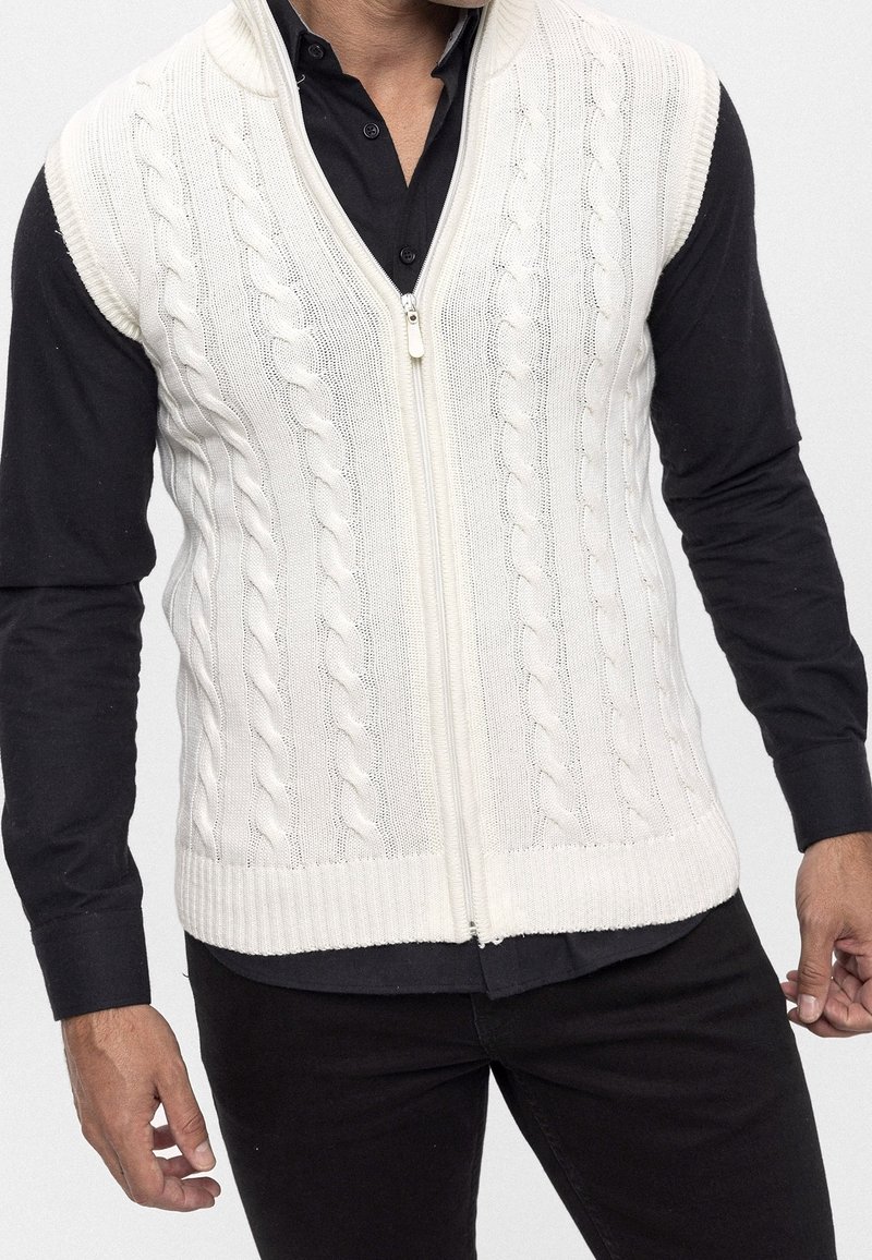 Kabelgebreid, crème mouwloos vest met een ritssluiting aan de voorkant; heeft een gestructureerd patroon en een geribde zoom; gedragen over een zwart shirt met lange mouwen.