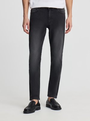 Uomo che indossa jeans neri slim-fit, camicia bianca infilata e mocassini in pelle neri, in piedi contro uno sfondo chiaro e uniforme.