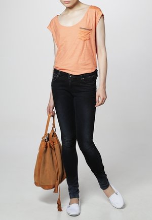 Oranžové volné tričko s kapsou, černé skinny džíny, bílé slip-on boty a béžová semišová taška na stažení s třásněmi.