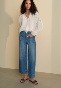 Blusa blanca de botones con tela texturizada, combinada con jeans de pierna ancha de talle alto en azul. La modelo lleva sandalias de estampado animal en blanco y negro.