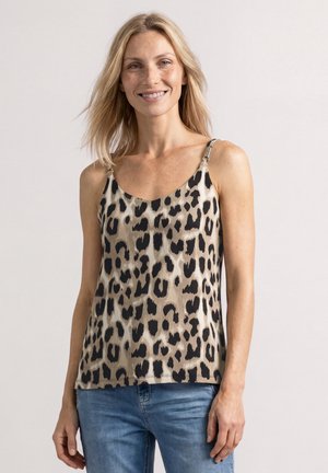 Smilende kvinde med blondt hår iført en ærmeløs beige top med sort leopardprint og blå jeans, stående foran en ensfarvet baggrund.