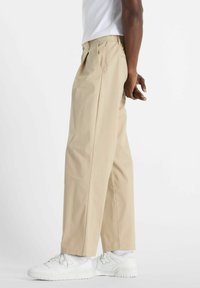 Beige trousers with a relaxed fit and pleated front. Paired with white sneakers, showcasing a smooth texture and classic design.