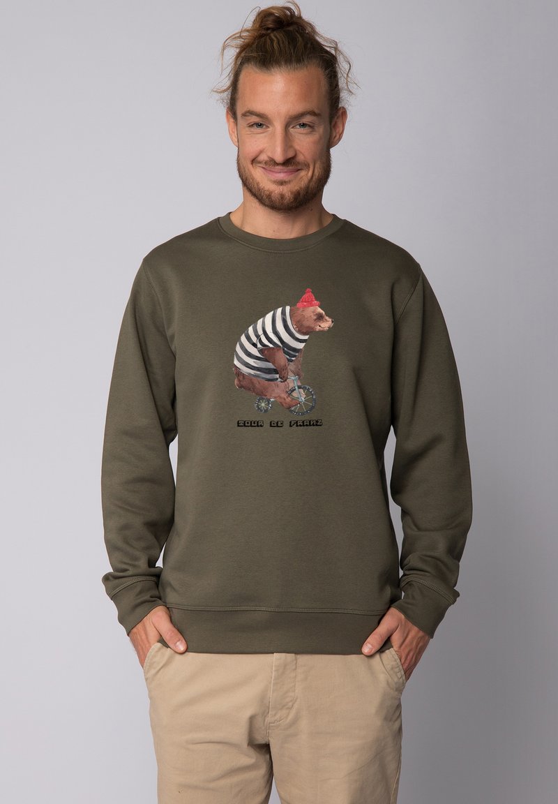 Olijfgroene sweatshirt met lange mouwen en een ronde hals. Heeft een illustratie van een beer met een rode hoed die op een fiets rijdt, vergezeld van tekst.