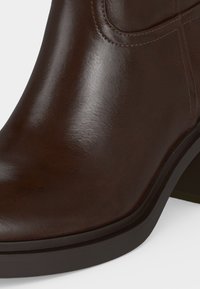 Gros plan sur une botte cheville en cuir lisse marron foncé avec semelle noire et talon bloc bas.