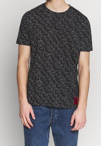 Sort t-shirt med et geometrisk hvidt mønster, rund hals, korte ærmer og et rødt mærke ved sømmen. Lavet af bomuldsstoff, afslappet pasform.