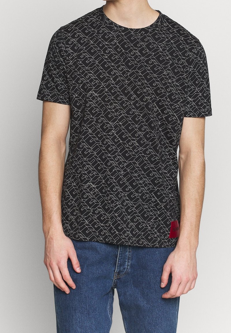Sort t-shirt med et geometrisk hvidt mønster, rund hals, korte ærmer og et rødt mærke ved sømmen. Lavet af bomuldsstoff, afslappet pasform.