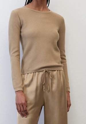 Personne portant un haut beige texturé à manches longues et un pantalon satiné beige assorti avec cordon, debout contre un mur clair uni.