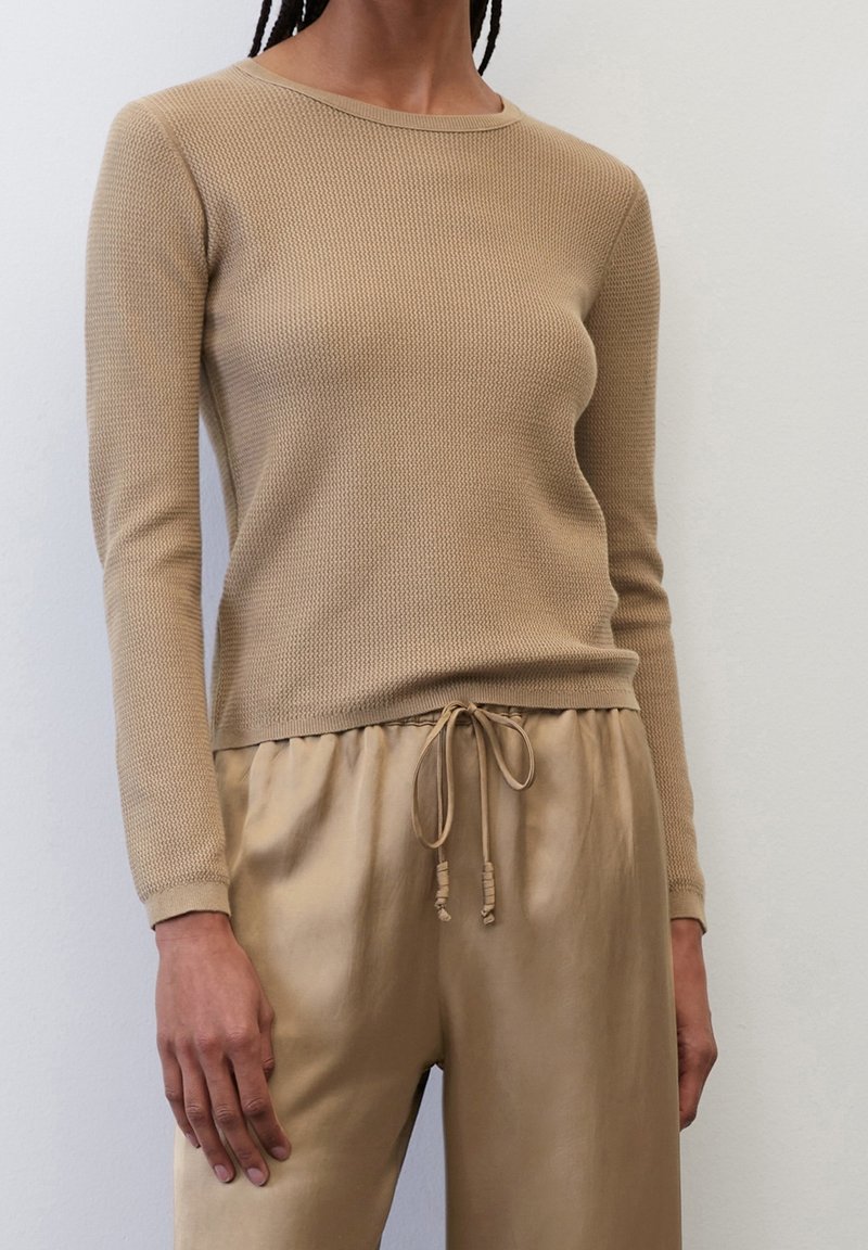 Personne portant un haut beige texturé à manches longues et un pantalon satiné beige assorti avec cordon, debout contre un mur clair uni.