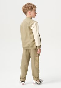 Khaki twee-delige set bestaande uit een beige langemouwen top en bijpassende joggers met een cargozak. Zwarte en witte ingebouwde schoenen met een ruitpatroon.
