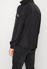 Czarna pikowana kurtka z wysokim kołnierzem, ściągaczami przy mankietach i bocznymi kieszeniami. Na lewej rękawie znajduje się logo CK Jeans. Gładka tekstura.