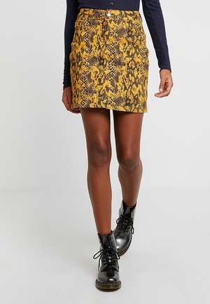 Mini-jupe à imprimé serpent jaune avec un design ajusté, présentant une fermeture à boutons sur le devant et une texture subtile. Portée avec des bottes noires à lacets.