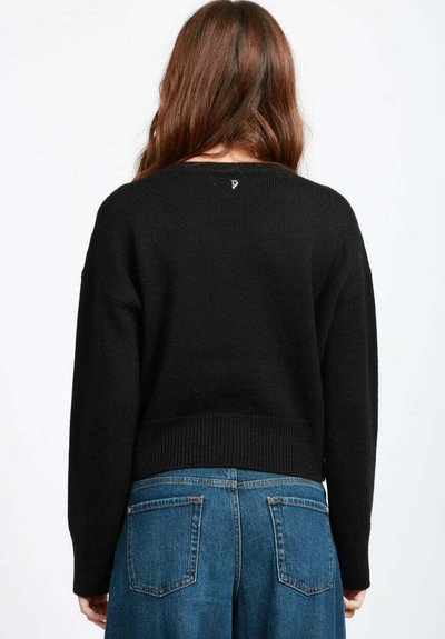 Donna con lunghi capelli castani che indossa un maglione nero a maniche lunghe e jeans blu, in piedi con la schiena rivolta verso l'osservatore su uno sfondo semplice.