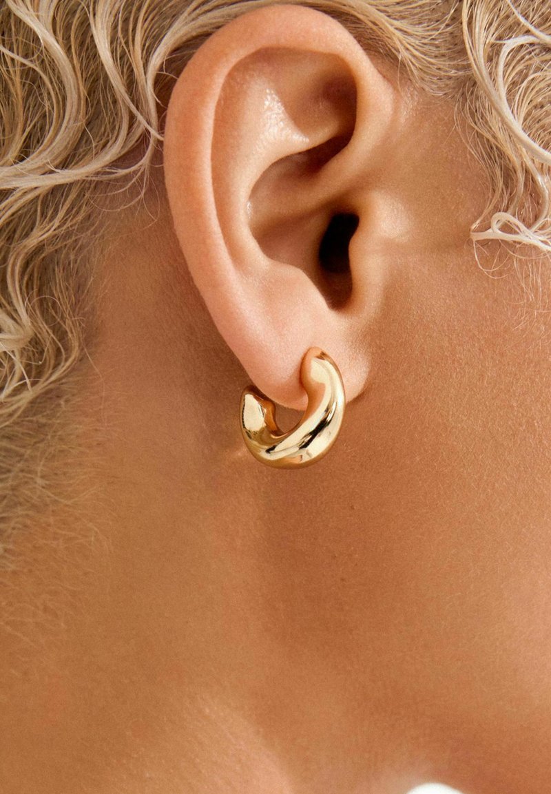 Zelta stīpas auskars ar gludu, spīdīgu virsmu, kuram ir asimetrisks dizains, kas ietver auss spraugu.