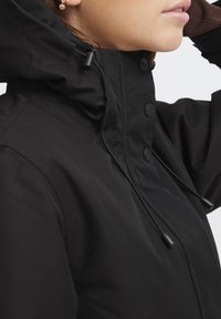 Veste à capuche noire avec un col haut, des boutons, un cordon de serrage réglable et une texture lisse. Accent sur la fonctionnalité et la protection contre les éléments.