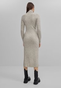 Robe-pull beige à manches longues avec col montant, silhouette ajustée et tissu en maille texturée. Associée à des bottines noires.