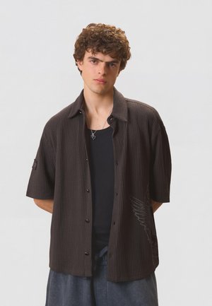 Jeune homme aux cheveux bouclés portant une chemise courte à manches courtes foncée et texturée ouverte sur un haut noir, debout les mains derrière le dos sur un fond uni.