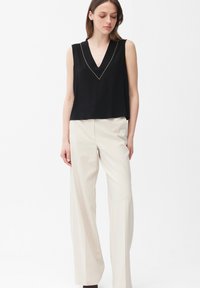 Mujer con top negro sin mangas de cuello en V con ribete dorado y pantalones anchos de talle alto color crema, de pie contra un fondo blanco.