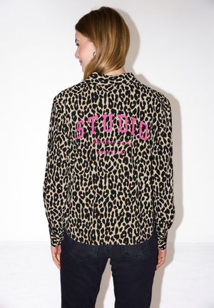 Femme aux cheveux châtain clair portant une blouse imprimée léopard avec le texte rose "STUDIO" au dos, assortie d'un pantalon noir, vue de dos.