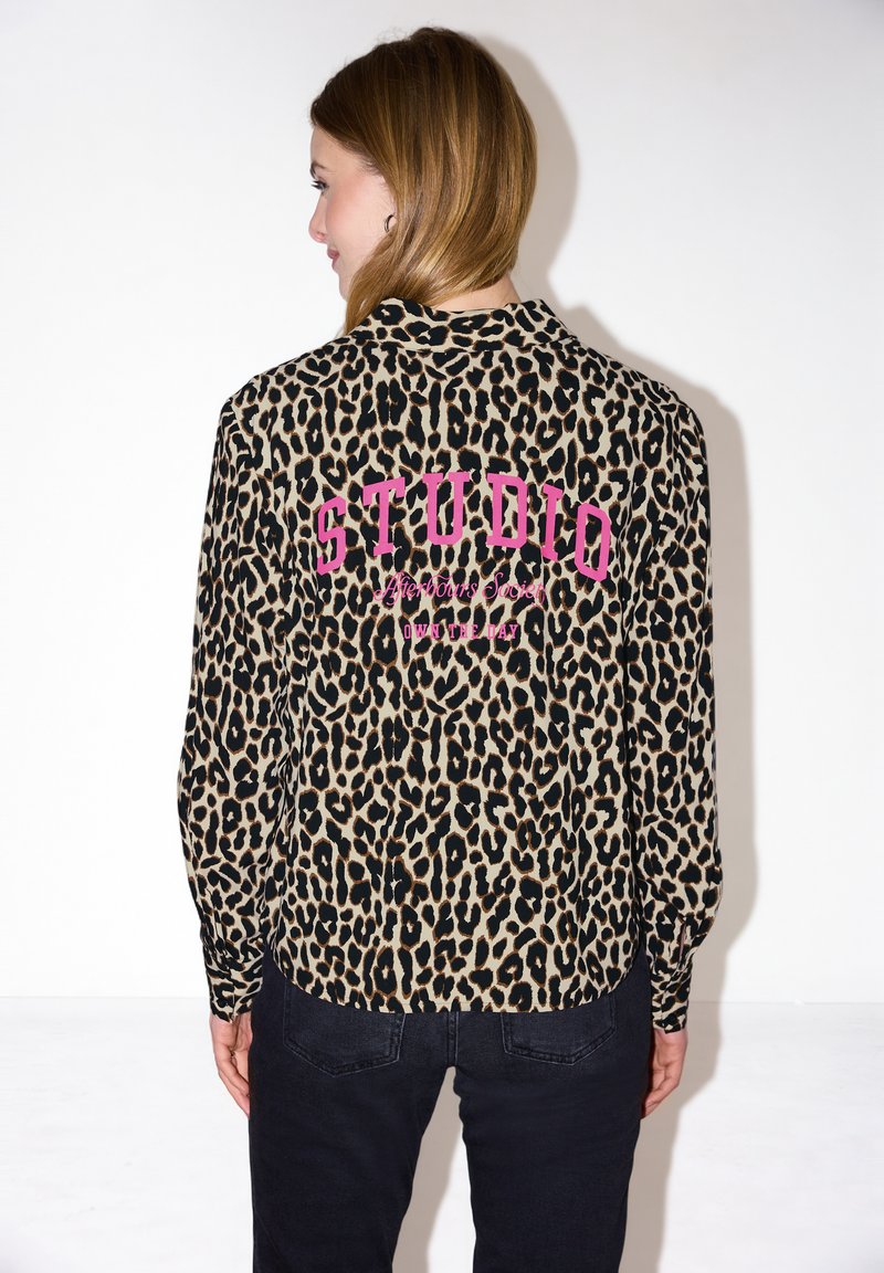 Frau mit hellbraunen Haaren, die eine Leopardenmuster-Bluse mit pinkem "STUDIO"-Text auf der Rückseite trägt, kombiniert mit einer schwarzen Hose, und von hinten zur Kamera gewandt ist.