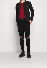 Chaqueta de mezclilla negra sobre una camisa roja, combinada con jeans skinny negros desgastados y botas negras. El material es tela, con detalles desgastados.