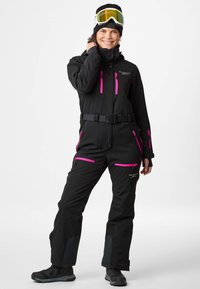 Black snowsuit with pink zippers and belt, featuring multiple pockets and a hood. Paired with a black beanie and ski goggles.