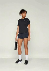 Mustad lühikeste varrukatega treeningpluus ja tumedad püksid, koos mustade tenniste ja valgete sokkidega. Modell hoiab käes musta veepudelit.