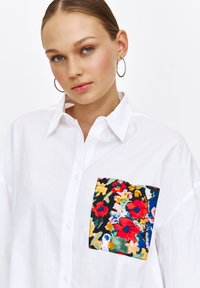 Biała koszula z guzikami z kolorowym wzorem kieszeni w kwiaty. Kieszeń zdobiona czerwonymi, żółtymi i niebieskimi kwiatami na ciemnym tle. Gładka faktura.