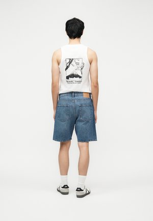 Uomo che indossa una maglietta bianca senza maniche con stampa del Monte Fuji e pantaloncini di jeans blu, in piedi di fronte a uno sfondo bianco uniforme.