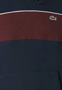 Hanorac bleumarin cu o dunga orizontală bordo, cu detalii albe și un logo de crocodil verde pe piept. Are un buzunar frontal.