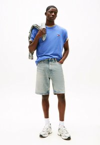 T-shirt di cotone blu con maniche arrotolate, pantaloni corti in denim chiaro e calzini bianchi abbinati a scarpe da ginnastica grigie e verdi. Giacca di jeans appoggiata sulla spalla.