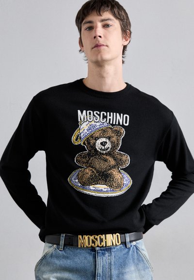 Pulover negru tricotat, cu un grafic de urs, farfurie circulară și logo-ul Moschino. Manechinul poartă blugi de denim albastru deschis, accentuați cu o curea aurie.