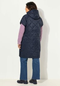 Námorně modrý prošívaný dlouhý vest s kapucí, krátké rukávy a diamantový vzor. Nositý přes světle růžový svetr a modré široké džíny. Černá obuv.