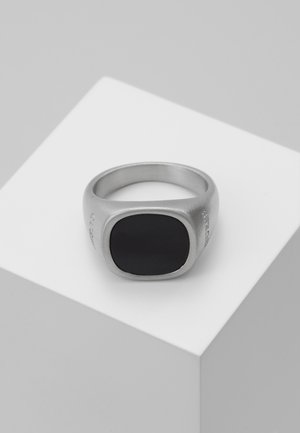 Bague chevalière en argent avec une pierre noire carrée et lisse au centre, présentée sur un socle blanc sur un fond gris clair.