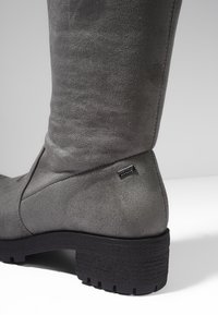 Botte grise en daim mi-mollet avec semelle noire texturée et petit talon carré, dotée d'une petite étiquette métallique rectangulaire de la marque près de la cheville.