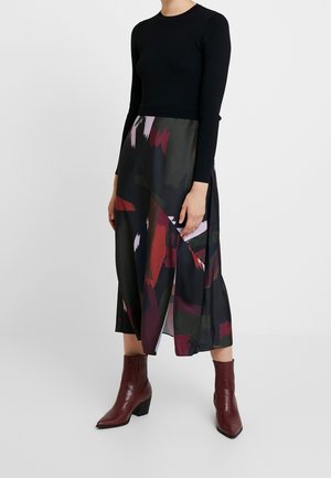 Kobieta ubrana w czarną bluzkę z długim rękawem, abstrakcyjną, wielokolorową spódnicę midi z bocznym rozporkiem oraz brązowe botki na słupku.