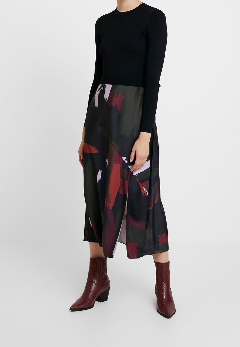 Femme portant un haut noir à manches longues, une jupe midi multicolore abstraite avec une fente latérale, et des bottines marron à talons blocs.