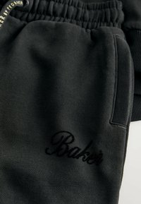 Primer plano de pantalones de chándal negros con cintura elástica, bolsillo lateral y texto bordado en negro que dice "Baker" en la tela.