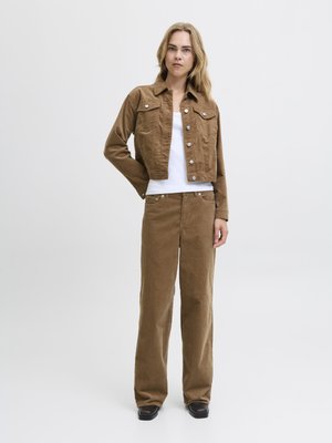 Bruine corduroyjas met voorzakken, gecombineerd met bijpassende wijde broek. De outfit heeft een witte tanktop eronder. Zwarte schoenen.